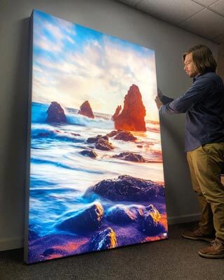 A man looking at a beautiful backlit wall art hanging of a beach and seaside rocks