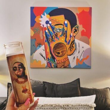 Massive colorful mac miller hip hop art in square hangs on white wall above grey couch in modern living room. A person holds a tall celebrity prayer candle of Mac Miller in front of the artwork, in tribute to the artist.