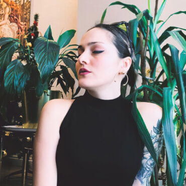 Woman artist Burcu poses with her chin down and to the side for a photo in front of indoor houseplants.