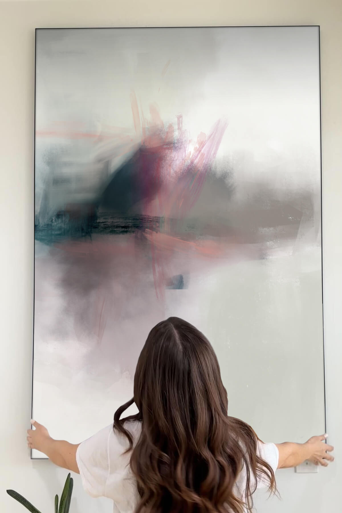 a girl with dark brown hair is hanging a pink and blue abstract artwork titled "Ephral" by Karine Tonial Grimm on a white wall in a modern living room. The extra-large abstract artwork features light grey background with pillowy brushstrokes in dark grey, dark teal, and light pink, adding a soft and muted touch to the room's decor.