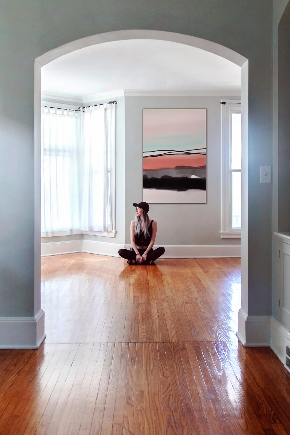 A girl sits on the floor in an empty blue living room, looking thoughtfully off into the distance while below a pink and blue abstract artwork with black accents, titled "Pink Horizon". The walls of the living room are painted light blue and the floor is a shiny walnut-colored wood, creating a serene and calming atmosphere in the room.