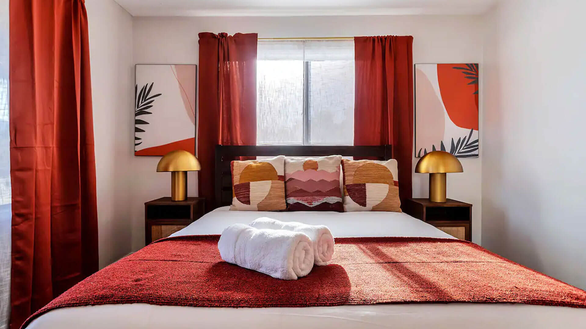 Queen size bed positioned in front of bedroom windows is decorated with matching bedside tables and contemporary lamps with complimentory abstract prints hanging above.