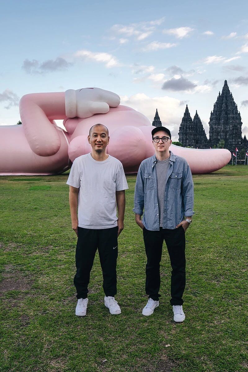 KAWS:HOLIDAY scuplture laying in the grassy lawn beside 9th century Indonesian temple, Prambanan Temple. KAWS and AllRightsReserved standing infront,