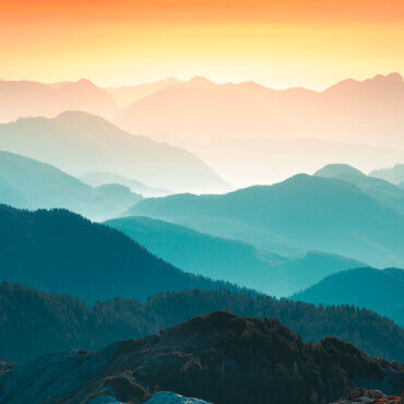 large mountain range, with light and dark blue hues, set in the foreground against a yellow sunrise.