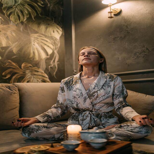 woman in a light blue robe meditating on a couch with a piece of floral wall art in the background