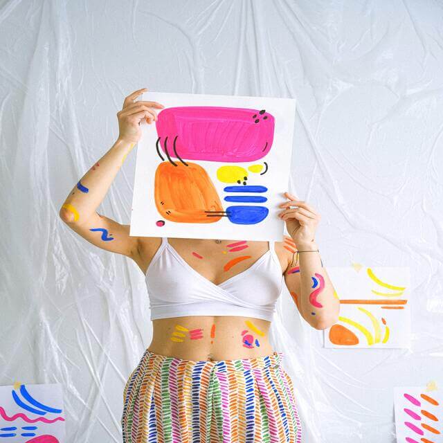 woman with paint on her clothes that is holding up a piece of colorful minimal art that she put together