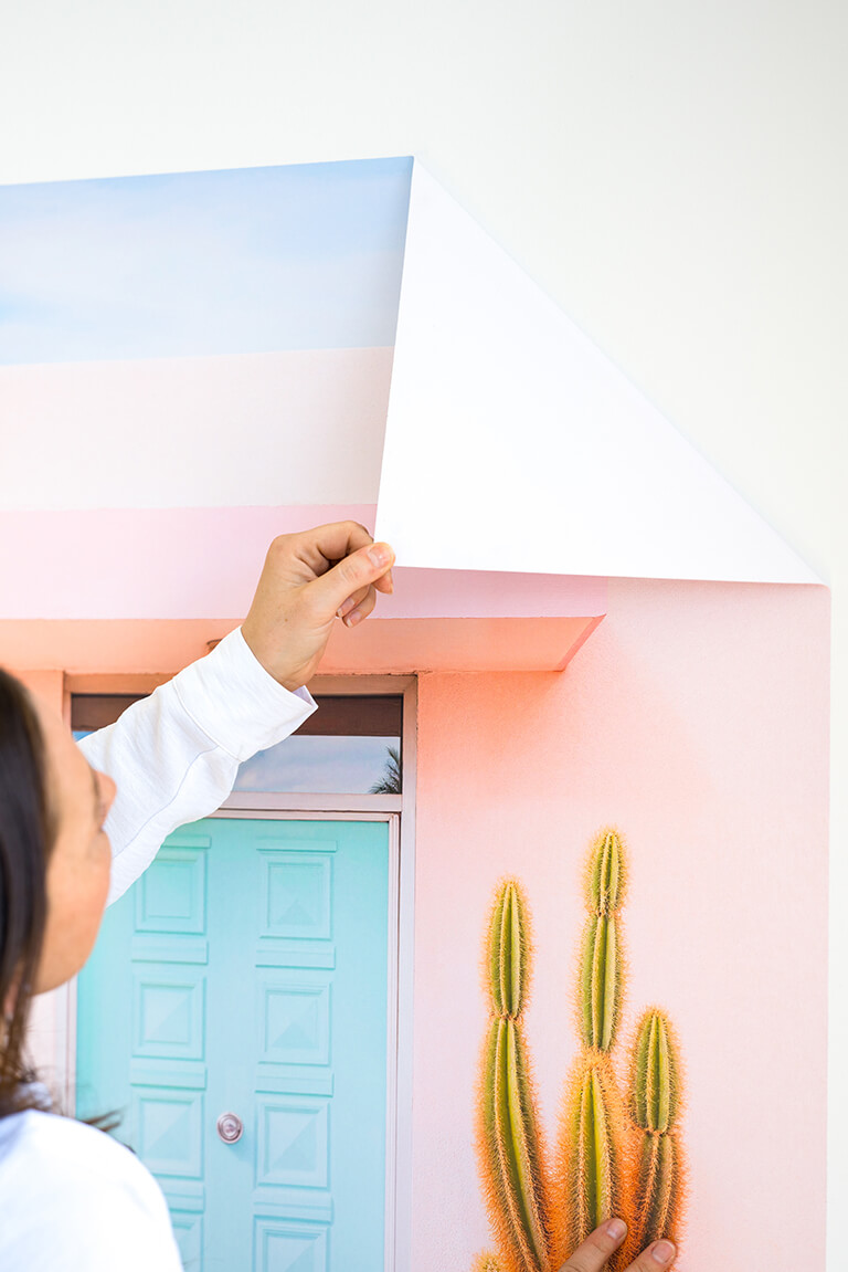 Person peeling back the corner of a large Stickablez wall art featuring a cactus and a pastel-colored door, showing the adhesive backing." Person peeling back the corner of a large Stickablez wall art featuring a cactus and a pastel-colored door, showing the adhesive backing." Person peeling back the corner of a large Stickablez wall art featuring a cactus and a pastel-colored door, showing the adhesive backing.