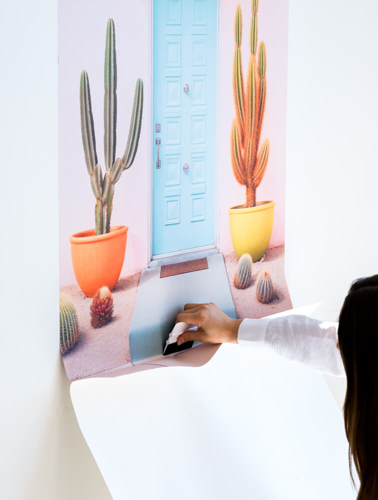 Person applying the lower part of a large Stickablez wall art featuring a cactus and a pastel-colored door, ensuring smooth adhesion.
