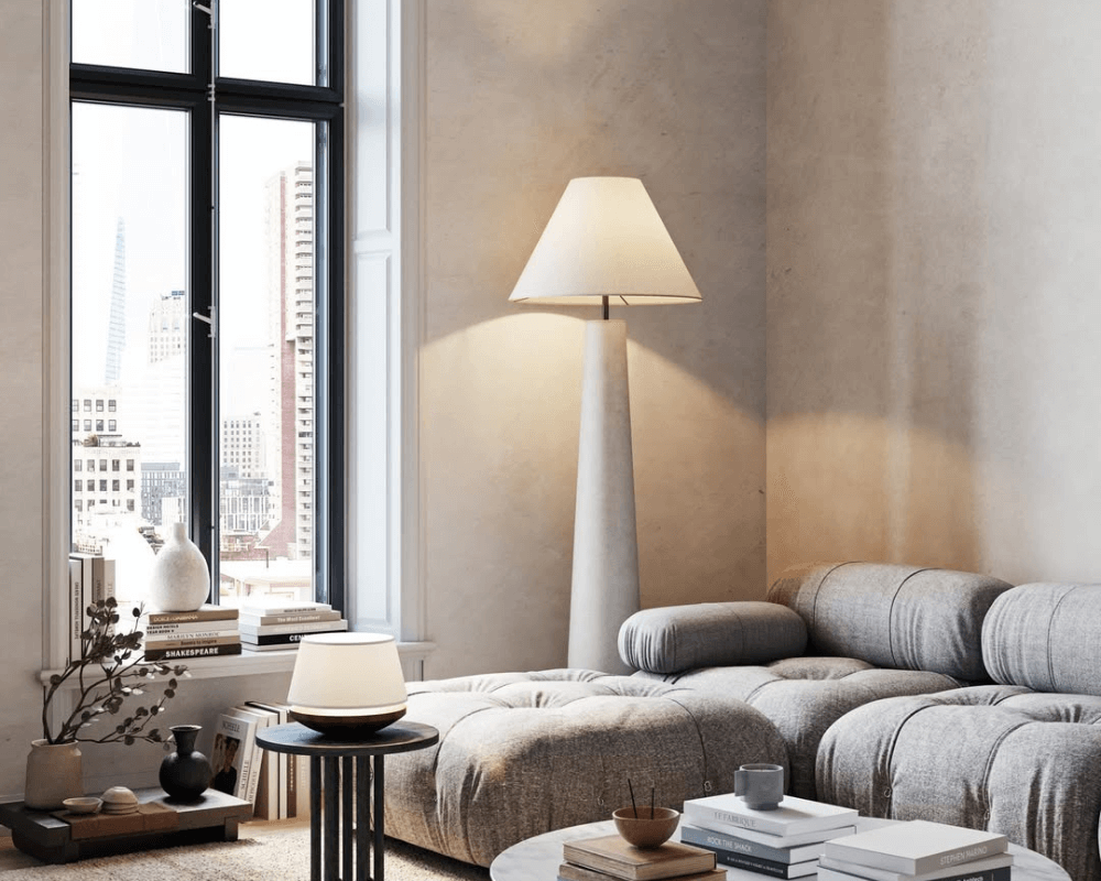A modern cement floor lamp with a sculptural design in a neutral-toned living room, next to a cozy gray sofa and large windows.