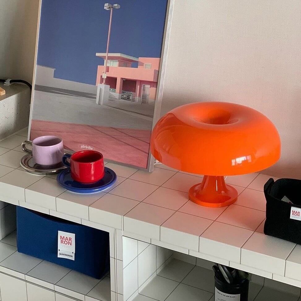 A bold orange mushroom-shaped lamp sits on a white-tiled shelf, accompanied by colorful coffee cups and a modern art print in the background.