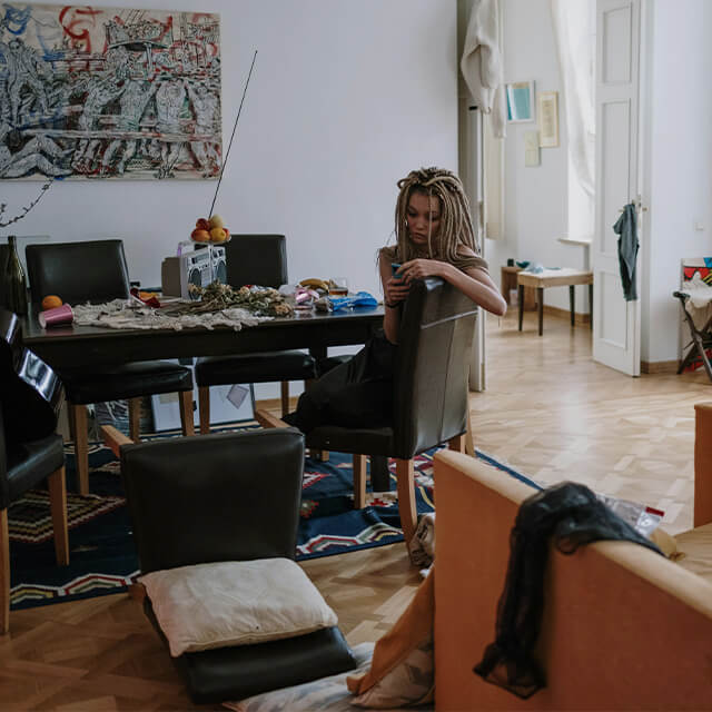 A woman sitting in a cluttered room with scattered items on the table and couch, creating a disorganized feel.