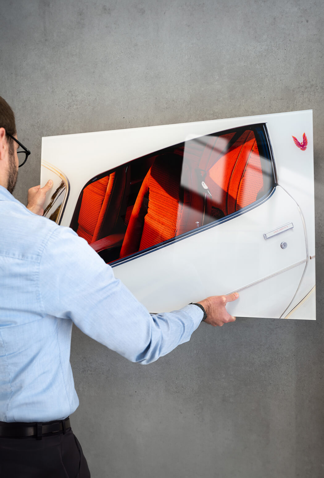 Man hanging a glossy acrylic print of a white car on a grey wall.