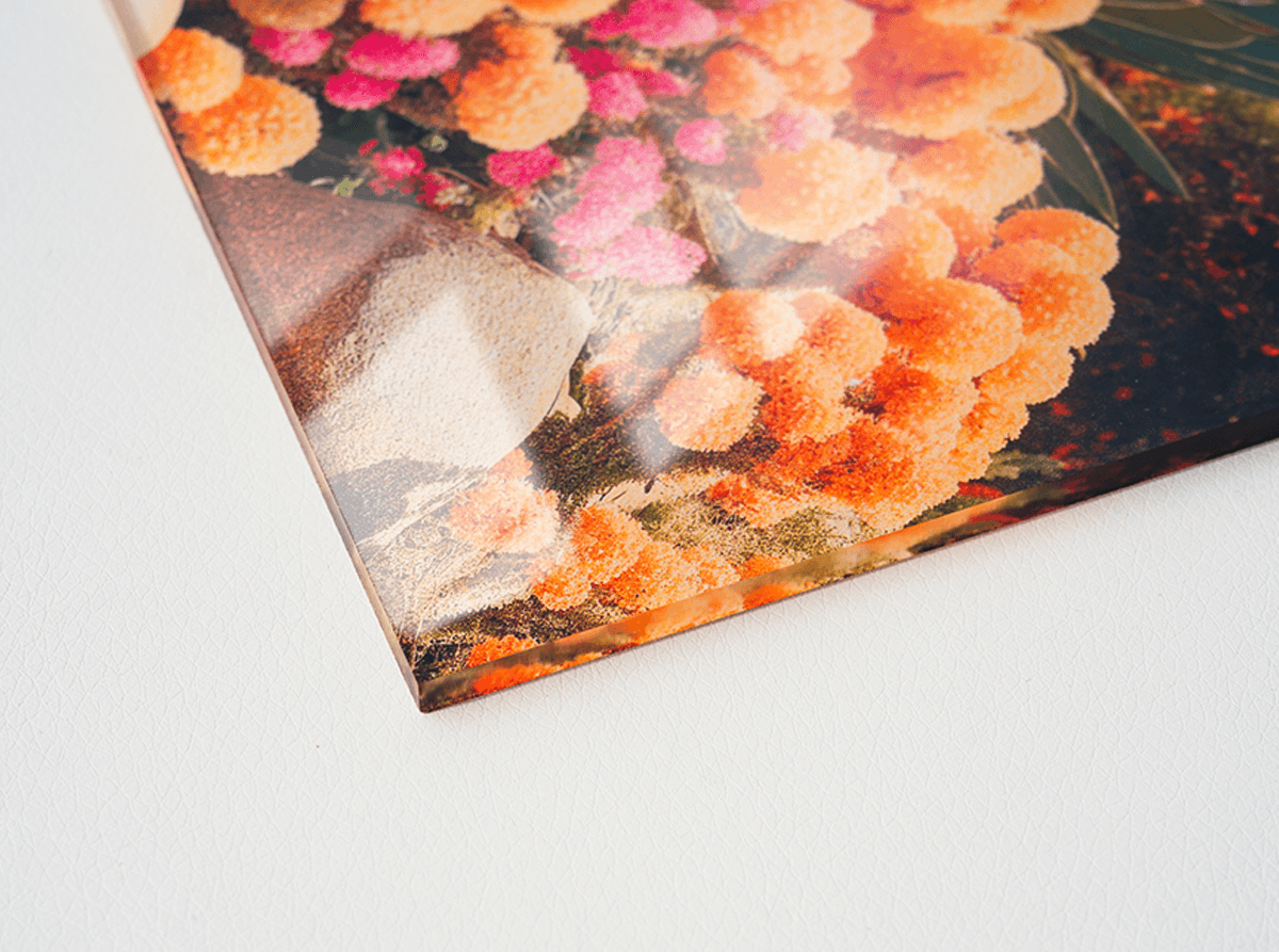 Close-up of an infused acrylic print corner showing glossy, glass-like depth with vibrant orange and pink flowers visible through the edge of the acrylic.