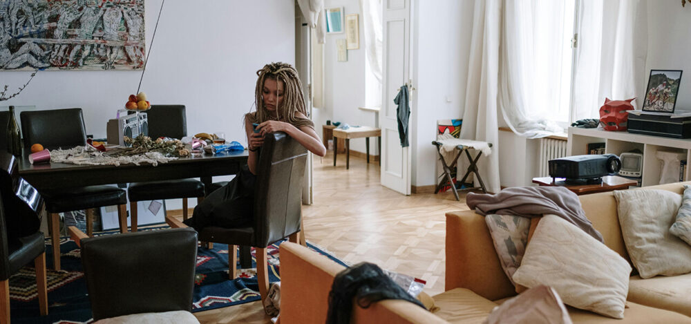 A woman sitting in a cluttered living room with scattered items on the dining table and couch, creating a messy and disorganized atmosphere.