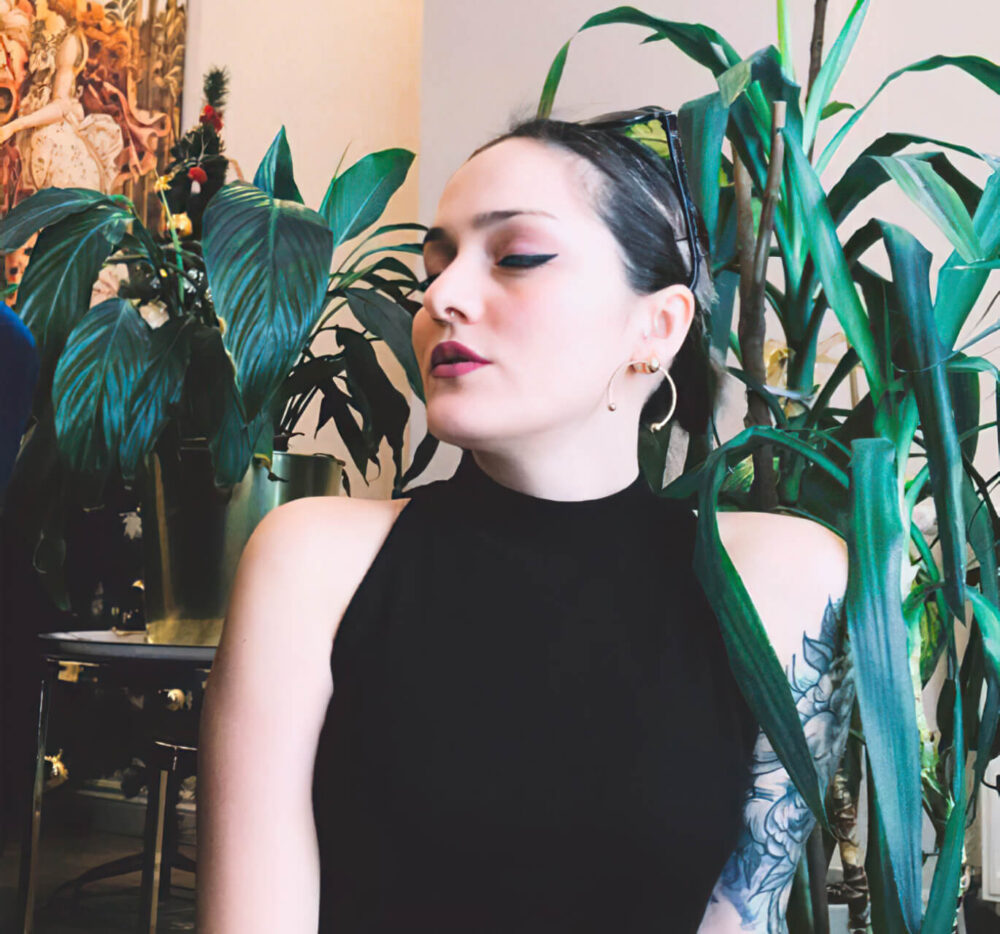 Woman artist Burcu poses with her chin down and to the side for a photo in front of indoor houseplants.