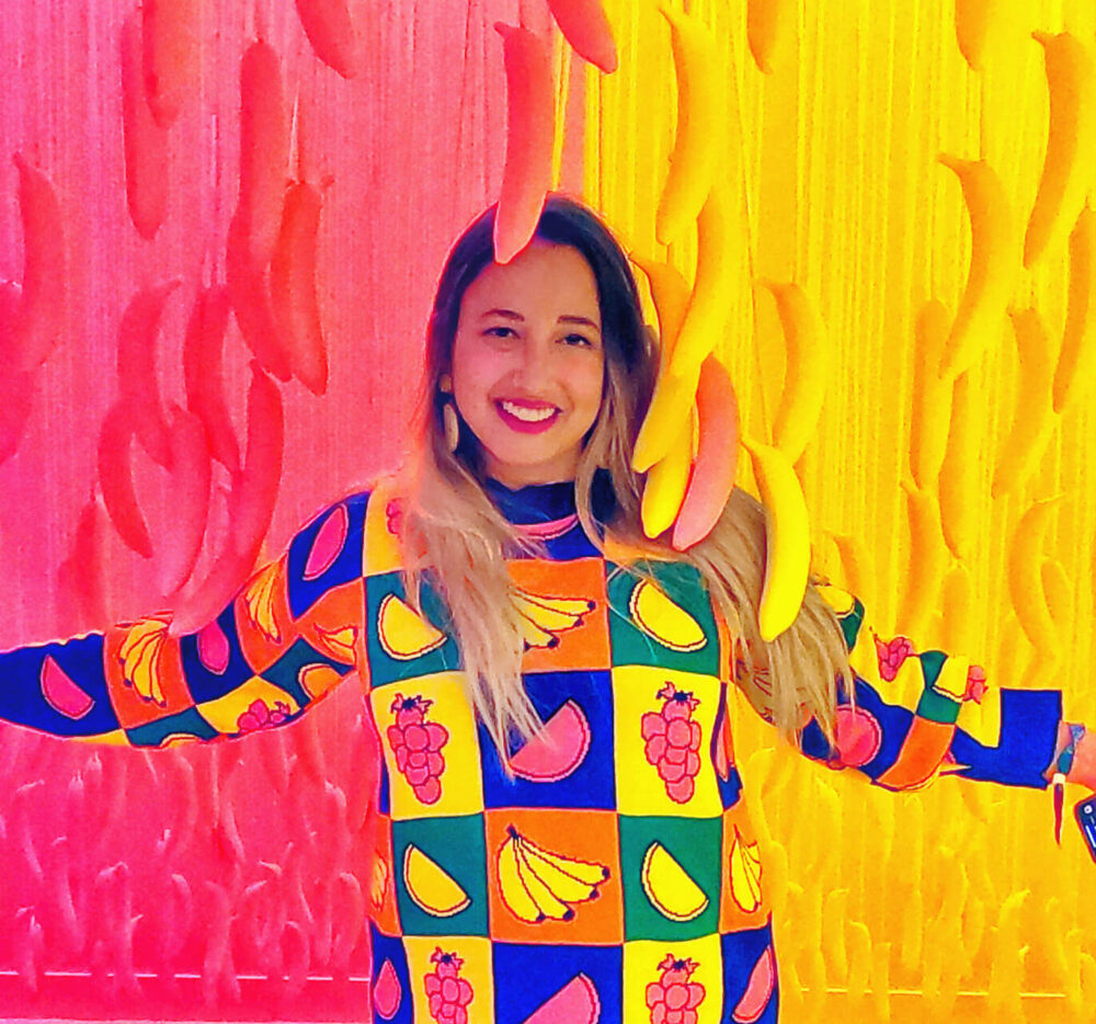 Female artist Jess Stempel poses in front of a pink and yellow backdrop with corresponding colored bananas hanging from the ceiling around her. She is wearing a colorful sweater and grins widely in this playful and creative setting.