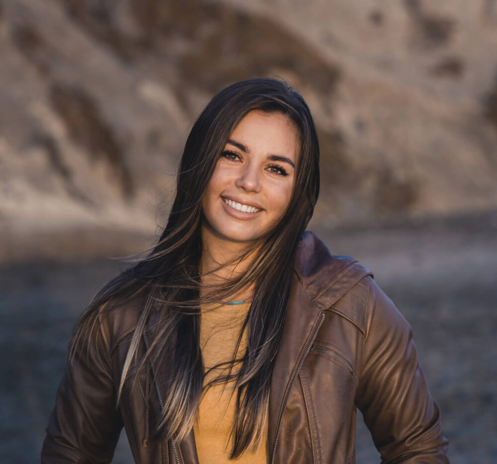Paige Tingey, a leading female artist making progress for women in Women's History Month, poses for a portrait in front of a deep blue lake at the side of a mountain, wearing a deep mustard yellow shirt below a brown leather jacket, and smiling as the wind gently blows her long brown hair.