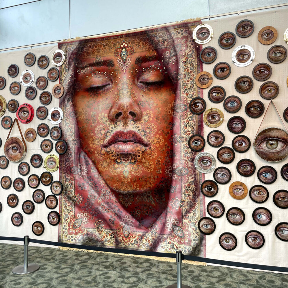 Massive rug hangs on the wall of the DeVos Place for ArtPrize 2022. The rug is painted with a women's face. She is wearing a hijab. Her eyes are closed, but surrounding the painted rug are small plates painted with open eyes.