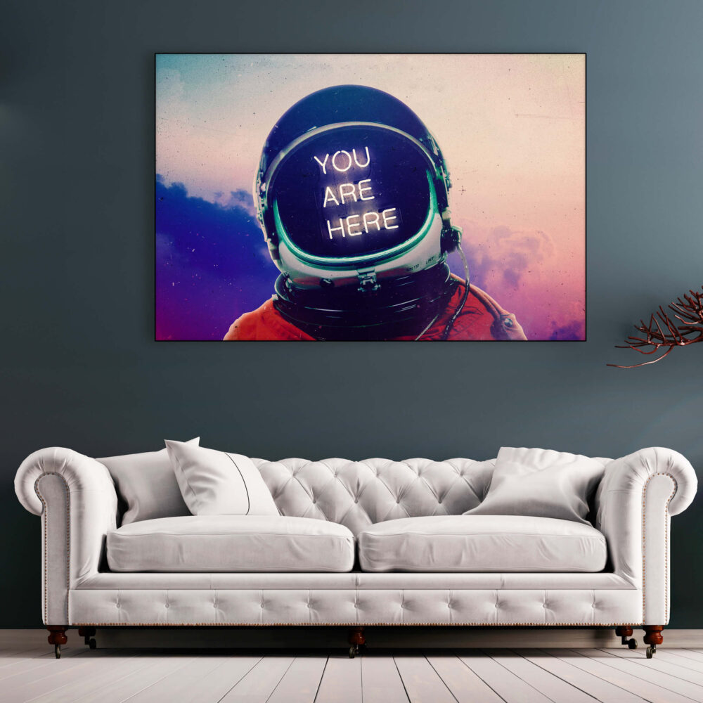 Large horizontal astronaut artwork with "you are here" in neon lettering hangs above neutral couch in living room