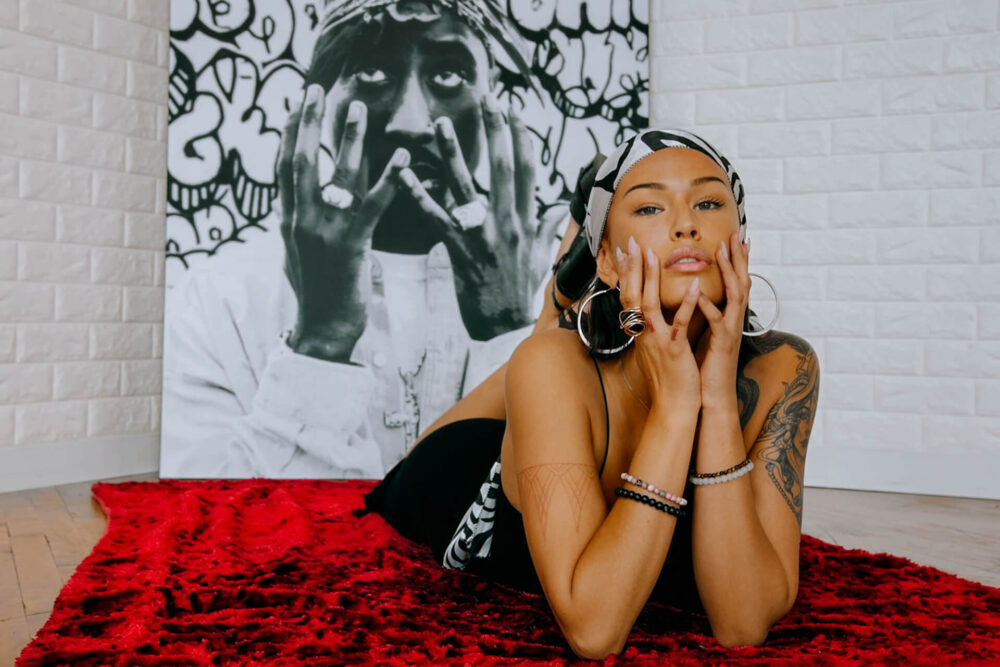 Large black and white photo of Tupac shows the rapper in distress, grabbing his face with both hands and looking up to the sky pleadingly. Behind Tupac the artist filled the background of the large artwork with black and white graffiti letters. The photo leans up on white painted brick walls. In front of the large wall art, a woman lays on a red blanket. She is propped up on her elbows and is gripping her face, mirroring how Tupac is grabbing his face in the photo behind her.