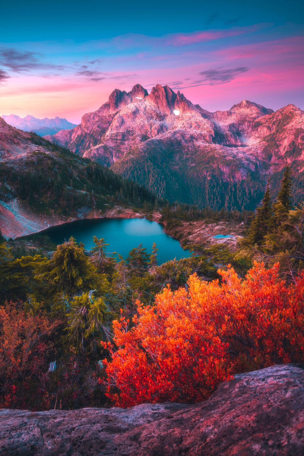 Colorful Alpine Mountain range with bright reds in the foreground, clear blue water in the midground and light pink sky in the background