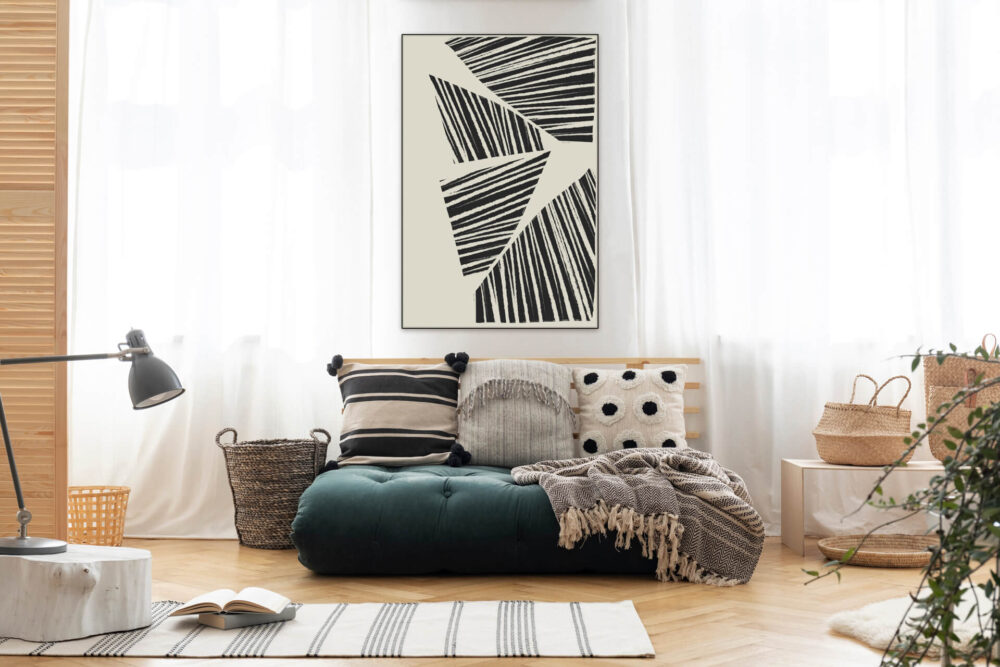 Black and white abstract striped art hangs on wall above boho couch between two windows in living room.
