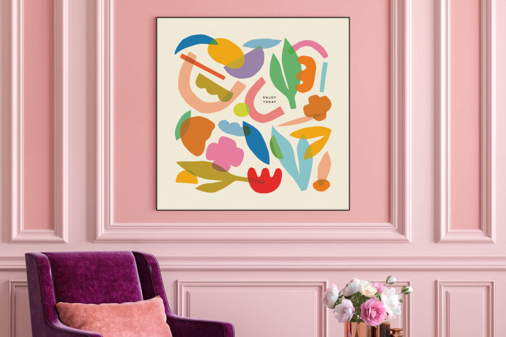Colorful wall art featuring abstract shapes layered over one another with the quote "Enjoy Today" written in black hangs on pink wall above dark pink velvet chair in living room.