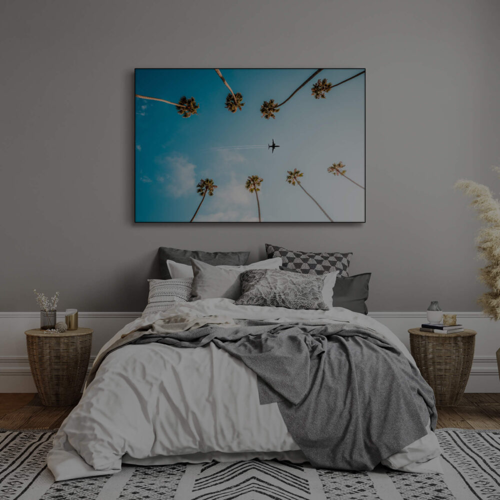 Photo featuring plane flying between palm trees through blue skies hangs above bed in bedroom decorated in neutral and grey.
