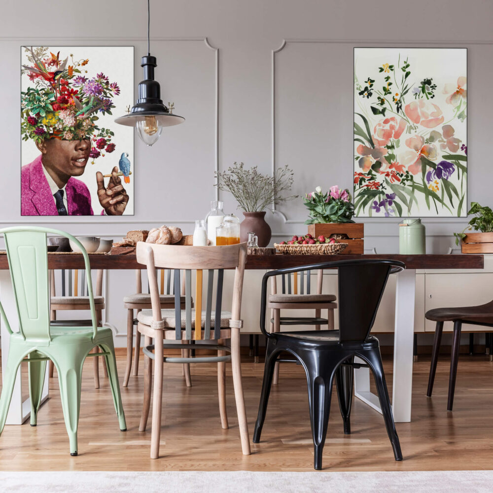 Maximalist dining room with flower wall art featuring "Igor" by BambashkART and "Floral Pink" by Shelby Kregel. Abundance of flowers, mis-matched chairs, colorful vases, and grey paneled walls with two large maximalist flower art pieces; one of a Black man in a pink jacket smoking with a little colorful bird perched on his hand, and the other of a wildflower bouquet in pink, green, and purple.