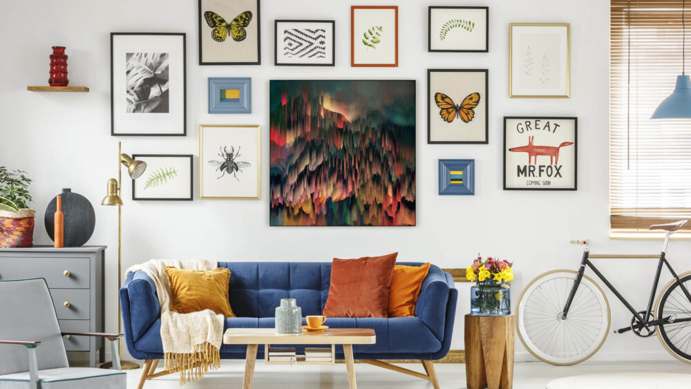 Maximalist living room with blue couch and gallery wall featuring a large colorful maximalist artwork in deep reds, yellows, and green, as well as small framed pieces featuring butterflies and insects on plain backgrounds. The artwork is the central focal point of the neutral-colored wall and makes the space feel energetic, while the blue velvet couch with orange and mustard yellow throw pillows completes the lively look.