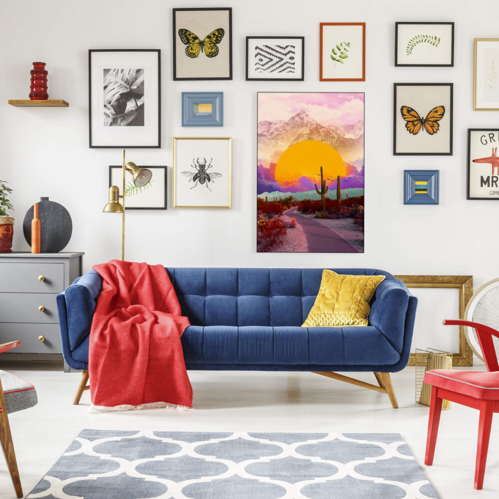 Maximalist living room with a boho gallery wall featuring "The Fallen Sun" by BambashkART, a large colorful maximalist art piece depicting the sun setting into clouds in front of a mountain in a surreal desert setting, as the focal point above a blue couch. The surrounding gallery wall includes framed prints of butterflies and other boho maximalist prints, showcasing the unique style of maximalist artists.