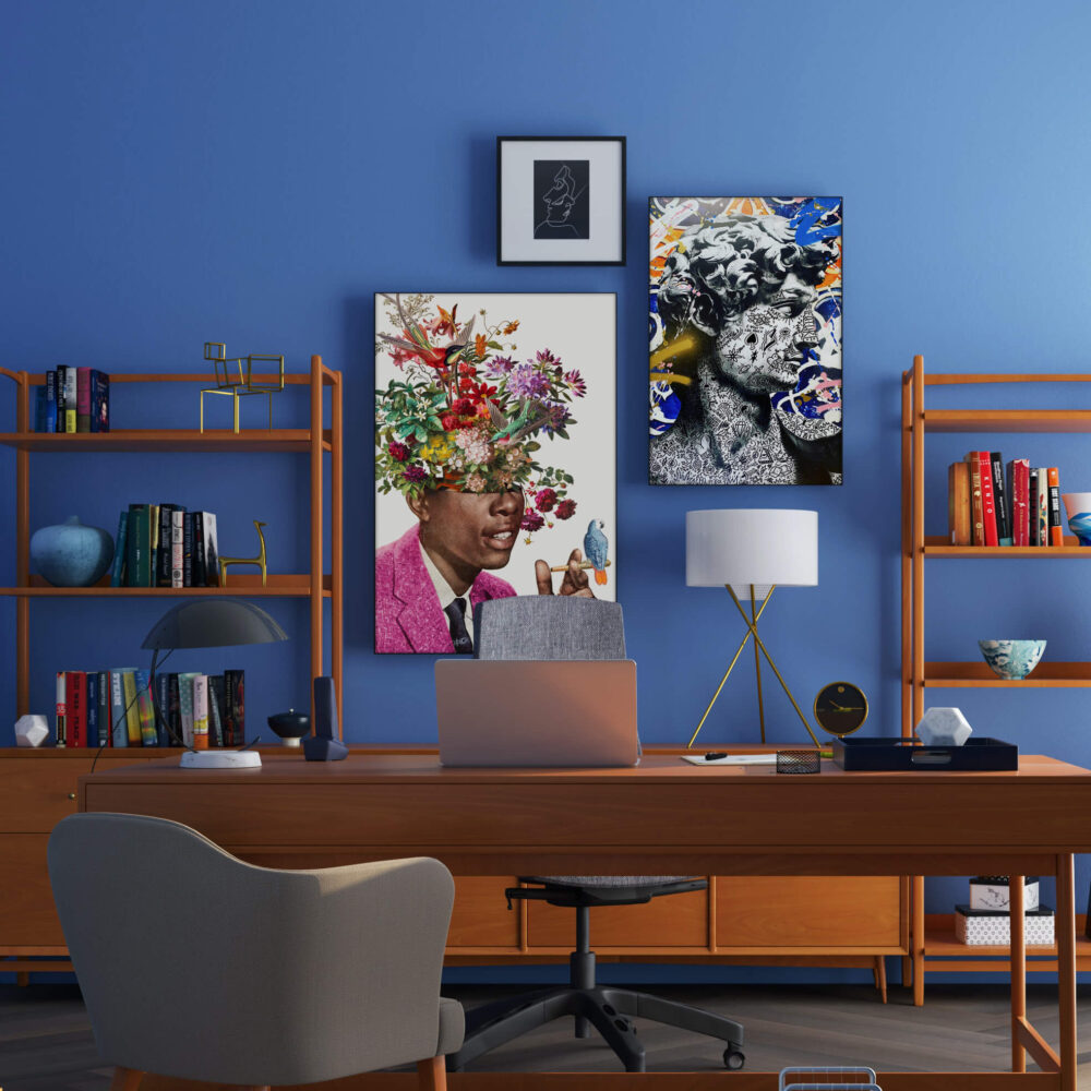 A bold and eclectic maximalist gallery wall is displayed in an office, featuring three large pieces of art hung on a bright blue wall. The art is complemented by decorative wooden shelves adorned with books and small trinkets, creating a unique and personalized aesthetic.