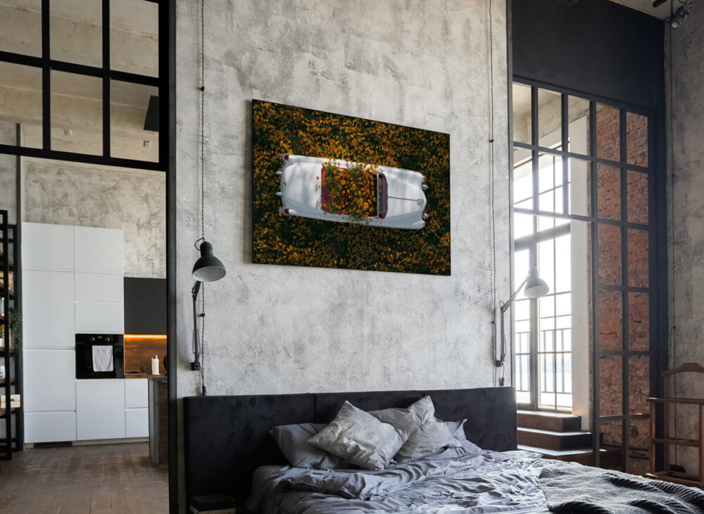 A large horizontal eclectic vintage artwork featuring a white antique Cadillac in a field of yellow flowers decorates a cement wall above the bed in a cool guy's bedroom with vintage eclectic décor. The bedroom is in a modern apartment building with exposed brick walls, tall ceilings, and massive windows, creating a unique and stylish living space.