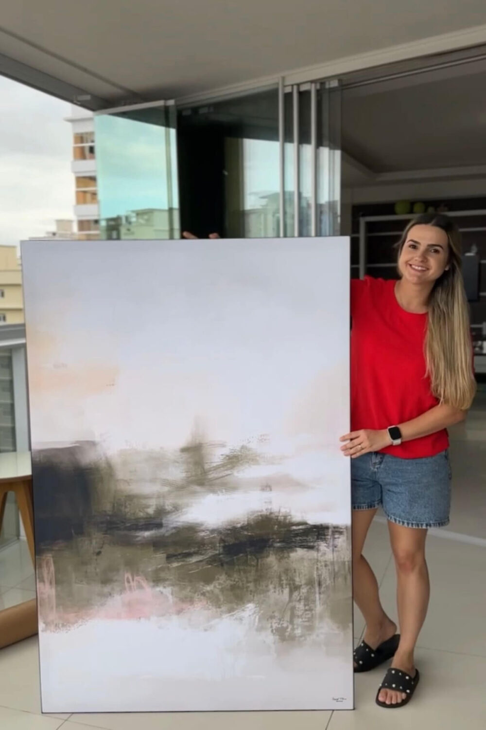 A young woman with long brown hair stands on an apartment balcony next to a massive abstract artwork in sage green and white. The artwork, featuring layers of sage green and soft pink, is almost as tall as the woman