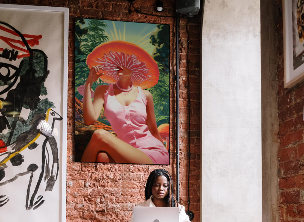 Large stoner wall art in pink, orange, and green features a woman with a mushroom for a head smoking. This cannabis inspired artwork hangs above a desk in an office or the desk in a dorm room
