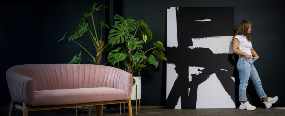 Large wall art leaned against the wall next to a woman, showing the vast size of the wall art. The wall art is a black and white abstract piece.