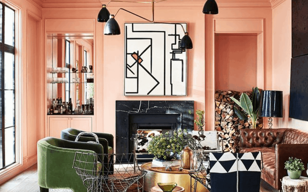 Colorful living room with pink walls, green velvet chairs, a brown leather sofa, and a black and white geometric artwork above a black fireplace.