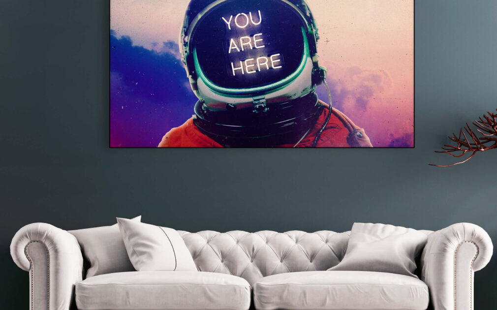 Large horizontal astronaut artwork with "you are here" in neon lettering hangs above neutral couch in living room