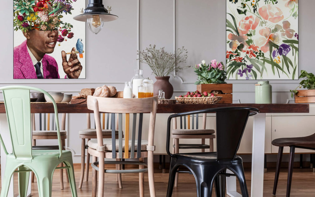 Maximalist dining room with flower wall art featuring "Igor" by BambashkART and "Floral Pink" by Shelby Kregel. Abundance of flowers, mis-matched chairs, colorful vases, and grey paneled walls with two large maximalist flower art pieces; one of a Black man in a pink jacket smoking with a little colorful bird perched on his hand, and the other of a wildflower bouquet in pink, green, and purple.