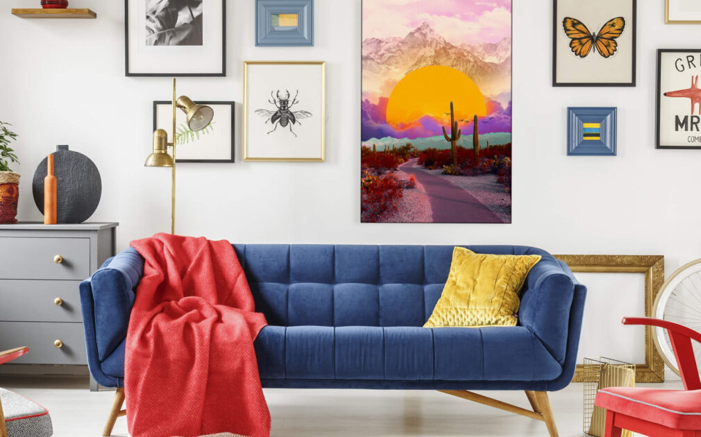 Maximalist living room with a boho gallery wall featuring "The Fallen Sun" by BambashkART, a large colorful maximalist art piece depicting the sun setting into clouds in front of a mountain in a surreal desert setting, as the focal point above a blue couch. The surrounding gallery wall includes framed prints of butterflies and other boho maximalist prints, showcasing the unique style of maximalist artists.