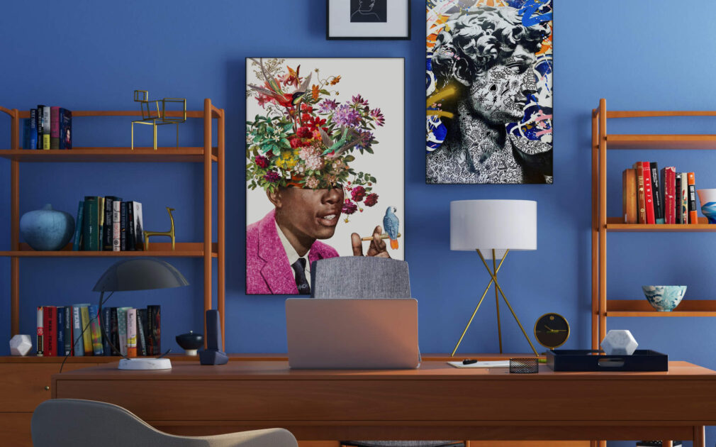 A bold and eclectic maximalist gallery wall is displayed in an office, featuring three large pieces of art hung on a bright blue wall. The art is complemented by decorative wooden shelves adorned with books and small trinkets, creating a unique and personalized aesthetic.