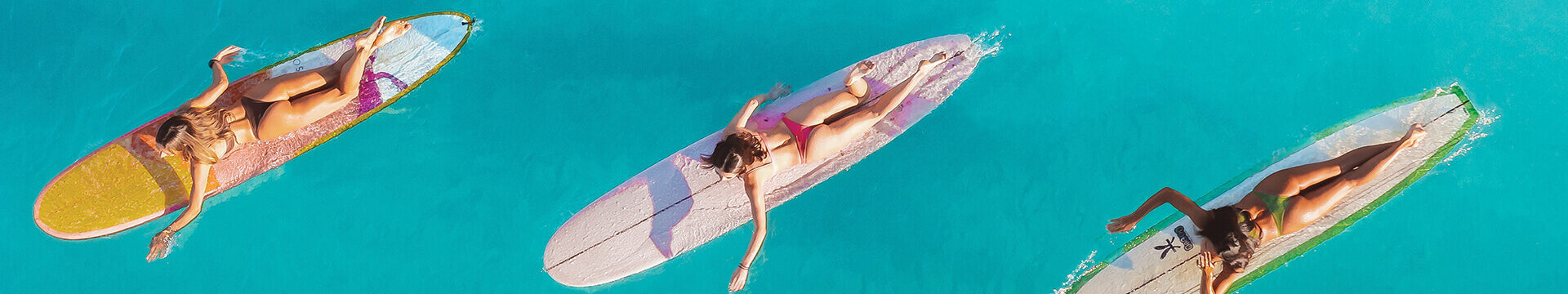 Photo of three girls swimming their surf boards out into the clear blue ocean.