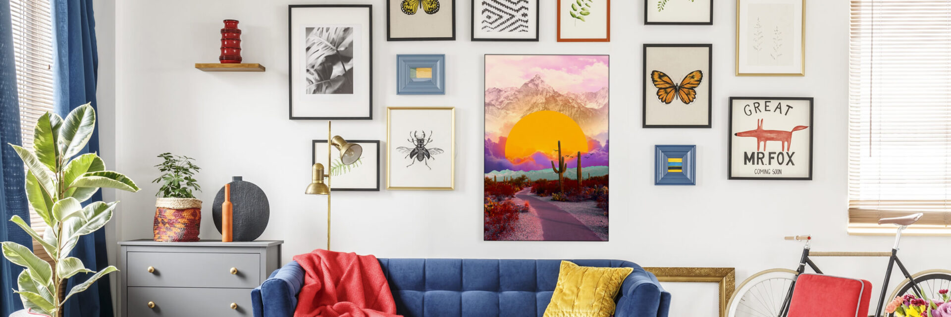 Maximalist living room with a boho gallery wall featuring "The Fallen Sun" by BambashkART, a large colorful maximalist art piece depicting the sun setting into clouds in front of a mountain in a surreal desert setting, as the focal point above a blue couch. The surrounding gallery wall includes framed prints of butterflies and other boho maximalist prints, showcasing the unique style of maximalist artists.