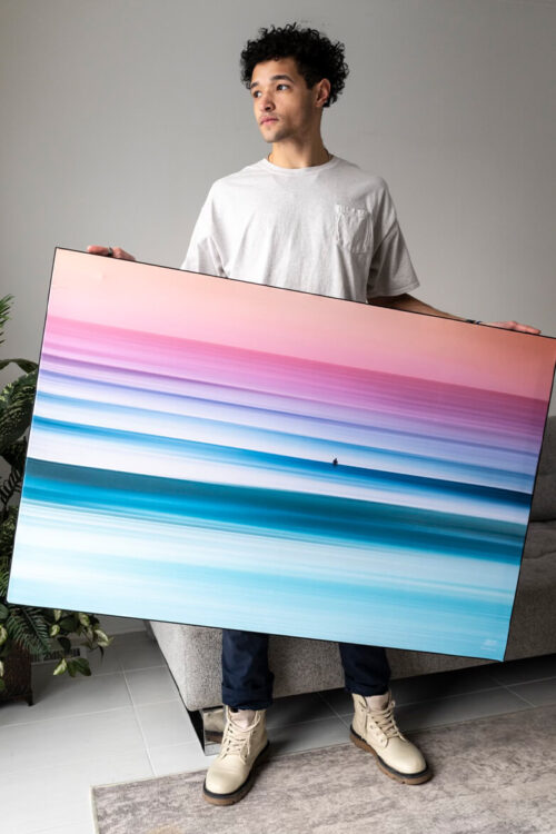 A black man wearing a white t-shirt, white boots, and blue pants is holding a large abstract sunset image in ombre pink to blue, while standing in a living room and looking off to the side.