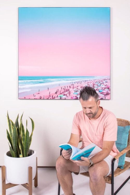 light pink and blue photo of the beach on a busy summer day hangs on a white wall in a living room above a man reading a magazine while sitting in a wicker chair.