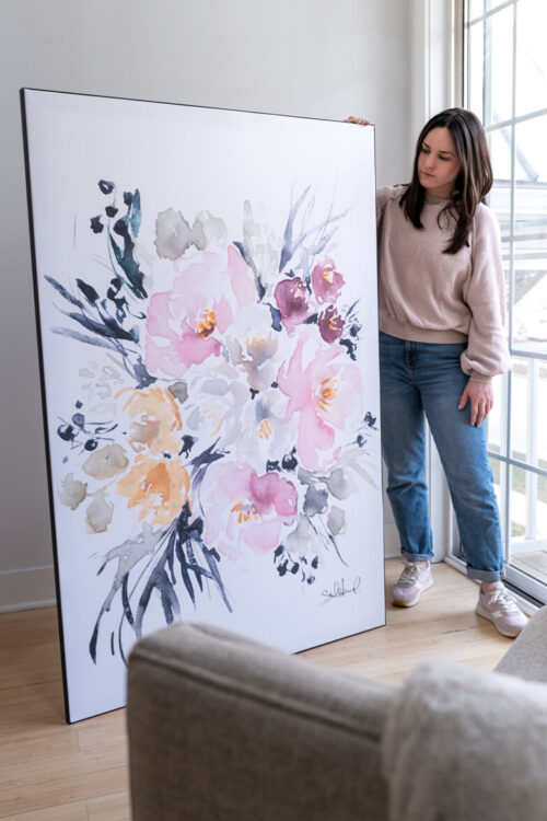 Girl wearing light pink sweater stands next to large watercolor painting of pink and yellow flowers in a neutral living room. The wall art is as tall as the girl holding the image.