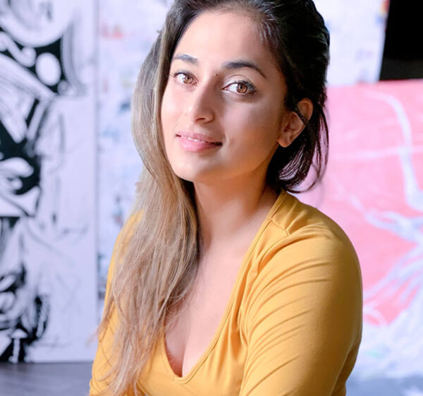 Female artist P Muir sits in her studio in front of a few of her paintings, wearing a yellow long sleeve shirt, and poses for Women's History Month.