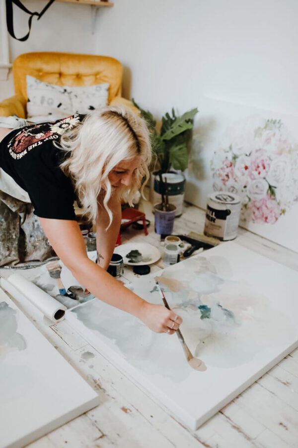 Female watercolor painter Shelby Kregel leans across a large canvas to paint in her studio, creating powerful yet feminine pieces in soft whimsical colors for Women's History Month.