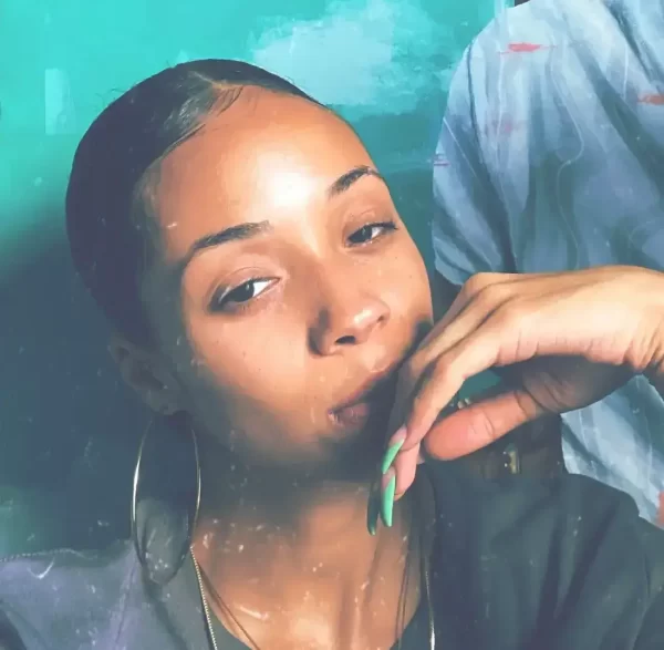 Black painter Kristen Campbell poses in front of a digital painting featuring a bright teal background and the back of a Black woman's head with curling rollers in her hair, wearing large gold hoop earrings and a sleek bun, for Women's History Month.