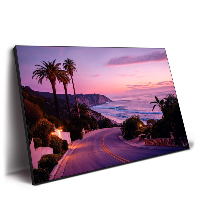 Beach road at sunset with palm trees, rugged cliffs, and a pink and purple sky.